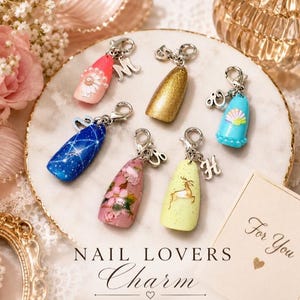 May include: Six decorative nail charms with silver lobster clasps and attached letter charms. Designs include a pink flower, gold glitter, blue shell, blue star pattern, pink floral, and yellow deer. The text reads NAIL LOVERS Charm For You.