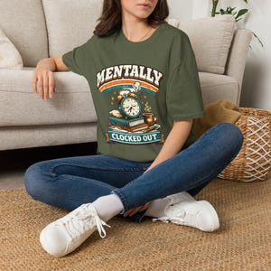 Peut inclure: Un t-shirt vert olive avec une impression graphique représentant un réveil, des livres et une tasse de café avec le texte MENTALLY CLOCKED OUT. Le motif inclut un arc-en-ciel rétro et des étoiles, porté avec un jean bleu et des baskets blanches.