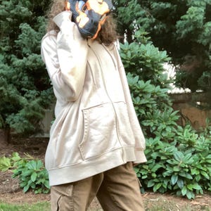 May include: A tan zip-up hoodie with a front pocket and brown cargo pants. A black and orange dinosaur mask is worn. The background features green foliage.