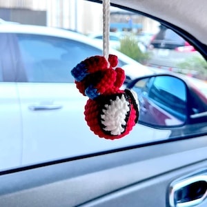 Könnte beinhalten: Ein handgefertigter Autoanhänger in Form eines Superheldenkopfes. Der Anhänger besteht aus roter, schwarzer, weißer und blauer Wolle und wird an einer weißen Schnur aufgehängt. Der Anhänger hängt im Inneren eines Autos.