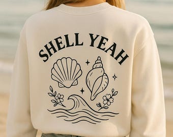 May include: Cream-colored sweatshirt with the words "SHELL YEAH" in black, along with seashell and wave graphics. The design includes a scallop shell, a conch shell, flowers, and stars. The model is wearing blue jeans.