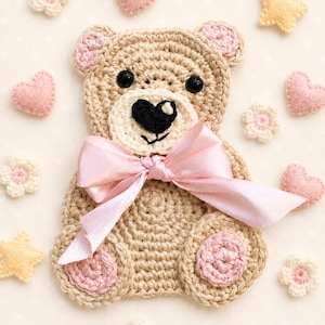 May include: A beige crochet teddy bear applique featuring a light pink satin ribbon bow at the neck, black plastic eyes, a black heart-shaped nose, and light pink circular accents on the ears and feet, surrounded by small felt hearts and flowers.