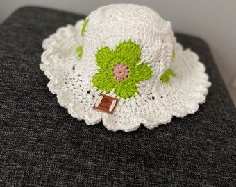 May include: A white crochet hat with a scalloped brim and a green and pink flower applique. The hat is handmade and appears to be for a child. The hat is sitting on a dark gray surface.
