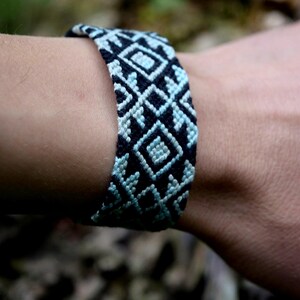 May include: A woven friendship bracelet featuring a repeating geometric pattern in black and light blue thread. The design consists of interconnected diamond shapes and angular lines, creating a textured, knotted appearance around the wrist.