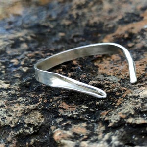 Op de afbeelding: Een zilveren manchetarmband met een gebogen, open ontwerp. De armband is gemaakt van een glanzend, metalen materiaal en heeft een gladde, gepolijste afwerking. De armband ligt op een ruw, getextureerd, donkerbruin en grijs stenen oppervlak.
