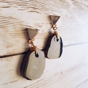 May include: A pair of silver and gold earrings with a unique, irregular, dark grey stone design. The earrings are hanging from a wooden surface.