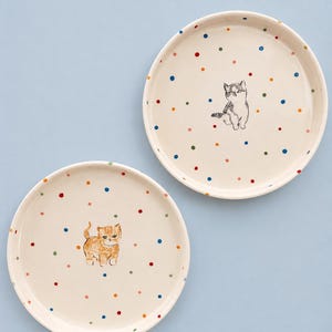 Può includere: Due piatti rotondi in ceramica con una base color crema caratterizzata da un motivo a piccoli pois multicolori. Un piatto mostra una piccola illustrazione di un gattino grigio e bianco, mentre l'altro presenta un piccolo gattino tigrato arancione.