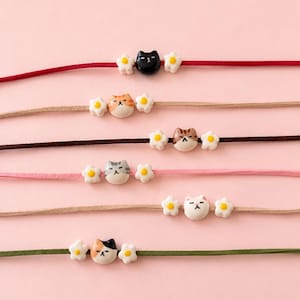 Può includere: Sei braccialetti con cordini in camoscio rosso, beige, marrone scuro, rosa, sabbia e verde oliva. Ogni braccialetto presenta una perla centrale in ceramica a forma di testa di gatto affiancata da due piccoli fiori bianchi con centro giallo.