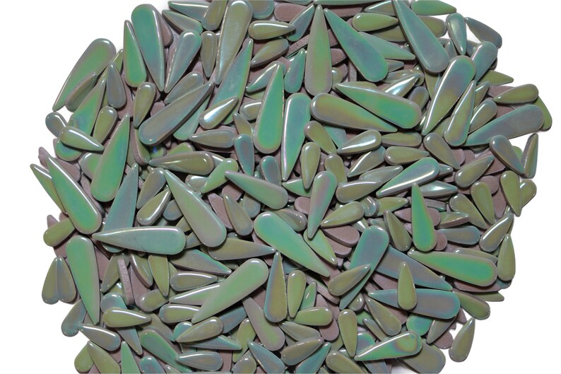 May include: A collection of iridescent, teardrop-shaped glass beads in shades of green, purple, and blue. The beads are scattered across a white background, creating a textured, organic pattern. These decorative beads are ideal for crafting and jewelry making.