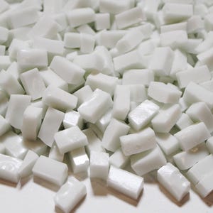 May include: A pile of small, rectangular, white mosaic tiles. The tiles are smooth and glossy, with slightly rounded edges. They are scattered in a random pattern, creating a textured surface. Ideal for craft projects.