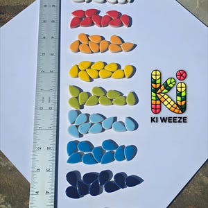 May include: A collection of colorful, teardrop-shaped decorative tiles arranged in rows. The colors include black, white, red, orange, yellow, green, light blue, dark blue, and purple. A ruler shows the tiles are approximately 1 inch in length. The logo "KI WEEZE" is also visible.