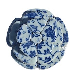 May include: A flower-shaped mosaic composed of white ceramic pieces with blue floral patterns. The mosaic is made up of irregularly shaped pieces, creating a textured, three-dimensional effect. The blue and white color scheme gives it a classic, elegant look.