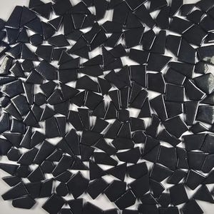 May include: An overhead shot displays a collection of irregularly shaped, glossy black tiles scattered across a white surface. The tiles vary in size and shape, creating a mosaic-like pattern. The lighting highlights the reflective quality of the tiles.