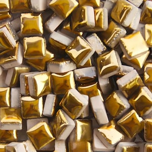 May include: Close-up of a collection of square mosaic tiles. The tiles have a gold, reflective surface on the top and sides, with a white base. The tiles are arranged in a random pattern, creating a textured surface.
