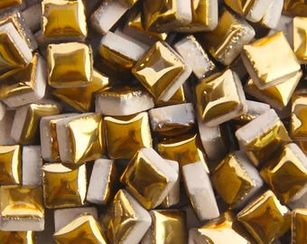 May include: Close-up of a collection of square mosaic tiles. The tiles have a gold, reflective surface on the top and sides, with a white base. The tiles are arranged in a random pattern, creating a textured surface.