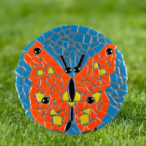 May include: A mosaic butterfly garden art piece. The butterfly design features orange, yellow, and black accents on a blue background. The mosaic is round and sits on a bed of green grass.