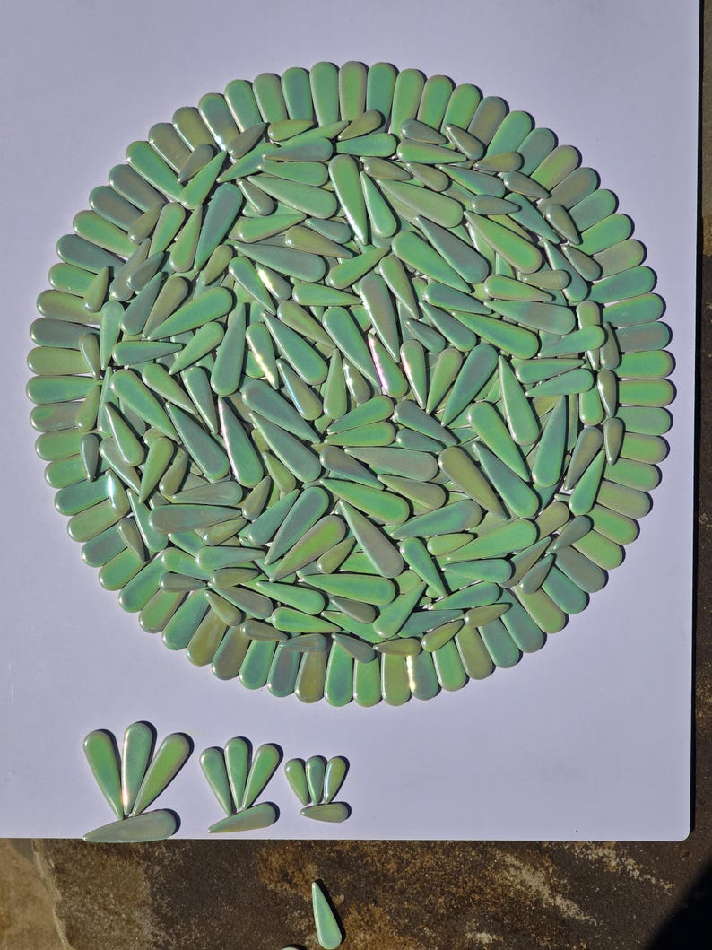 May include: A circular mosaic design composed of numerous green, teardrop-shaped tiles. The tiles are arranged in a radial pattern, with a border of similar tiles. Several smaller tile arrangements are below the main design.