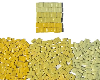 May include: A collection of small, square glass mosaic tiles in various shades of yellow and pale lime green. The tiles are arranged in a neat rectangular grid at the top, while a large assortment of loose tiles is scattered across the bottom area.