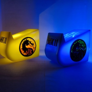 Könnte beinhalten: Zwei beleuchtete, stilisierte, gebogene Kästen. Einer leuchtet gelb mit einem Drachenlogo und dem Text "MK11", der andere blau mit einem grünen Logo und dem Text "MK11". Die Kästen stehen auf einer reflektierenden Oberfläche mit blauem Hintergrund.