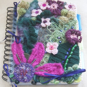 Op de afbeelding: Een handgemaakte journalhoes met een kleurrijk gehaakt ontwerp met bloemen, bladeren en kralen. De hoes is bevestigd aan een spiraalgebonden notitieboek.
