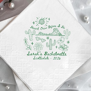 May include: White square paper napkins with a green desert-themed design. The design includes cacti, a mountain range, and the text "Desert Crew Before I Do" and "Sarah's Bachelorette Scottsdale - 2026". The napkins are folded and stacked.