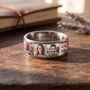 May include: A silver-toned metal band ring featuring a continuous wrap-around design with four distinct photographic portraits printed on the exterior surface, showcasing a couple, a woman, a man, and a baby in a clear, high-resolution format.