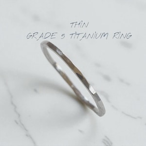 Könnte beinhalten: Ein dünner, silberfarbener Ring aus Titan Grad 5 mit einer glatten, polierten Oberfläche und einem einfachen, kreisförmigen Banddesign. Der Text "THIN GRADE 5 TITANIUM RING" ist über dem Ring deutlich sichtbar und gibt das Material an.