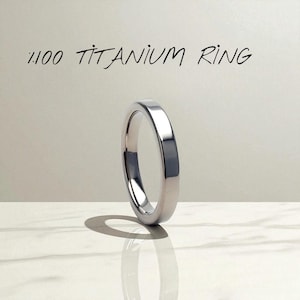 May include: A polished titanium ring with the text "1.100 TITANIUM RING" in black script. The ring is silver and sits on a white surface, casting a shadow. The ring is a simple band design.