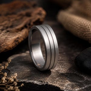 May include: A silver ring featuring three parallel grooves. The ring, crafted from brushed metal, rests on a textured stone surface. This simple yet elegant ring is a piece of jewellery.