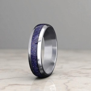 Op de afbeelding: Een zilveren ring met een donkerblauwe, glinsterende inleg. De ring heeft een gepolijste, metallic afwerking en een band van donkerblauw materiaal. De ring staat tegen een neutrale achtergrond.