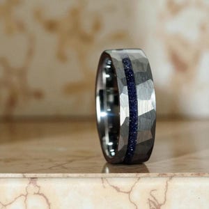 Puede incluir: Un anillo plateado con textura martillada y una incrustación azul oscuro y brillante. El anillo se muestra sobre un fondo neutro, resaltando su diseño único. La banda es gruesa y parece ser de metal.