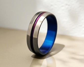 May include: A metallic ring featuring a smooth, brushed gray exterior surface with a thin, recessed purple groove running along the center. The interior of the band is finished in a vibrant, solid blue color, creating a distinct contrast.