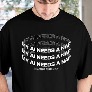 Könnte beinhalten: Schwarzes T-Shirt mit dem Text "MY AI NEEDS A NAP" in einem gebogenen Design. Der Satz "CHATTING SINCE 2025" ist darunter gedruckt. Das Shirt ist ein lässiger Unisex-Stil.