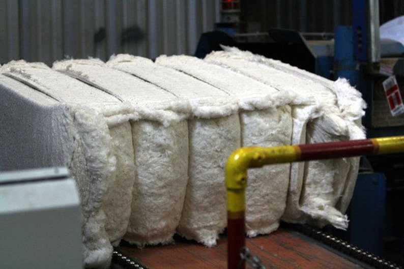 Puede incluir: Primer plano de una pila de fardos de algod&oacute;n blanco en una cinta transportadora en un entorno de f&aacute;brica. Los fardos est&aacute;n apretados y tienen una textura esponjosa.