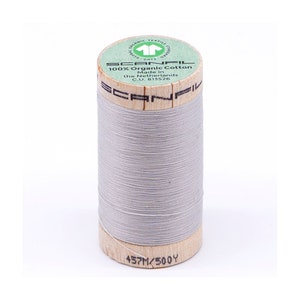 May include: A spool of light grey organic cotton thread with a wooden bobbin. The label reads "SCANFIL 100% Organic Cotton Made in the Netherlands C.U. 813526" and "457M/500Y".