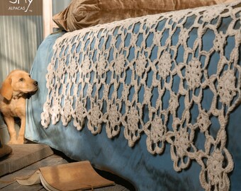 Puede incluir: Una cama azul con una manta de ganchillo blanca y una almohada de terciopelo marrón. La cama está en una habitación con suelo de madera y un perro está sentado en el suelo junto a la cama.