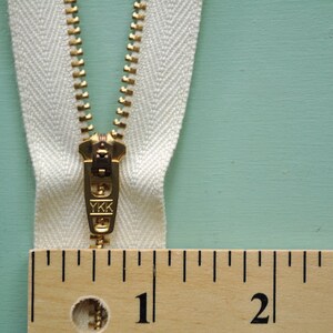 May include: Close-up of a cream-colored zipper with a gold-colored metal pull. The zipper is measured with a wooden ruler, showing the first two inches. The zipper pull is marked with the letters "YKK".