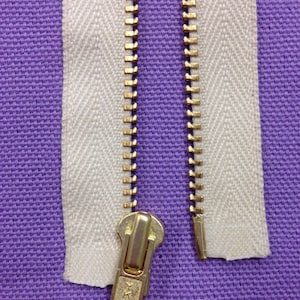 May include: A white zipper tape with a gold metal zipper pull and teeth. The zipper tape has a herringbone pattern.