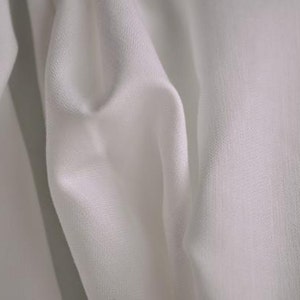 May include: Close-up of white fabric with a textured surface. The fabric is draped, creating soft folds and shadows. The material appears to be lightweight and possibly used for clothing or home decor.