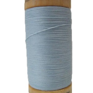 Puede incluir: Una bobina de hilo de algodón orgánico 100% de color azul claro con una bobina de madera. La bobina está etiquetada con "110Y / 100m" y "100% Algodón Orgánico".