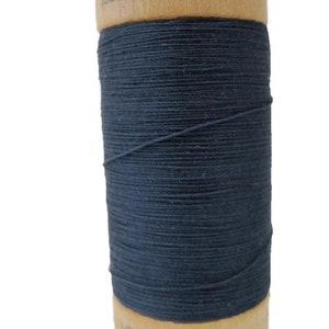 Puede incluir: Una bobina de hilo de algodón orgánico 100% azul oscuro en una bobina de madera. La bobina está etiquetada con "100% algodón orgánico" y "1000Y".