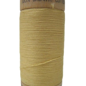 Puede incluir: Una bobina de hilo de algodón orgánico 100% en color amarillo claro. La bobina es de madera y tiene una etiqueta que dice "100% algodón orgánico".