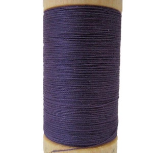Puede incluir: Una bobina de hilo de algodón orgánico morado en una bobina de madera. La bobina está etiquetada con "Organic Cotton".