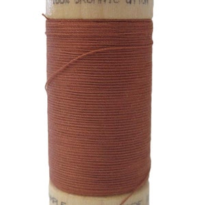 Puede incluir: Una bobina de hilo de algodón orgánico 100% en color terracota. La bobina es de madera y tiene el texto "100% ORGANIC COTTON" impreso en la parte superior.