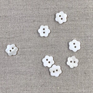 以下が含まれることがあります： 2つの穴がそれぞれ開いた白い花の形をしたボタンが7つ。ボタンは薄い灰色の布地に並べて置かれています。