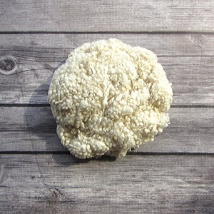 May include: A round, off-white, textured object with a dense, nubby surface. The item appears to be made of a soft, fibrous material, possibly wool or a similar textile. The object is set against a gray wooden background.