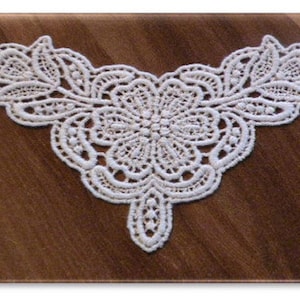 May include: A white lace applique with a floral and leaf design is centered on a rectangular wooden surface. The lace features intricate details and a delicate, openwork pattern. The wood has a warm, brown tone.