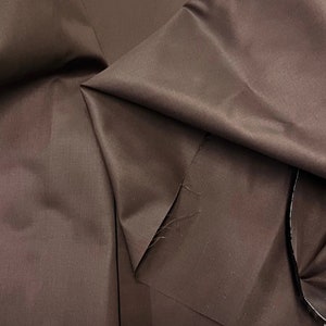 Puede incluir: Primer plano de una tela marrón con una textura suave y sedosa. La tela está doblada de forma que se aprecia su caída y brillo.