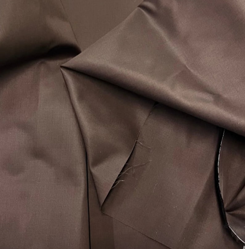 K&ouml;nnte beinhalten: Nahaufnahme eines braunen Stoffes mit glatter, seidenartiger Textur. Der Stoff ist so gefaltet, dass seine Falten und sein Glanz sichtbar werden.