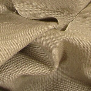 May include: Close-up of a beige fabric with a textured weave. The fabric is folded and wrinkled, showing the texture and drape.